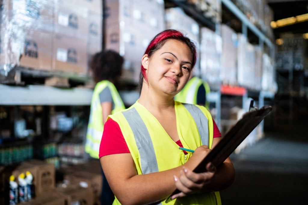 A photograph of a young woman holding a clipboard in a warehouse.