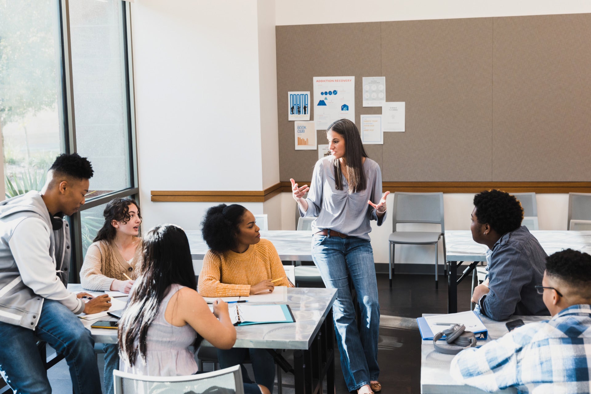 A photograph of a teacher leaning on a desk speaking to a group of high school students sitting around a table.