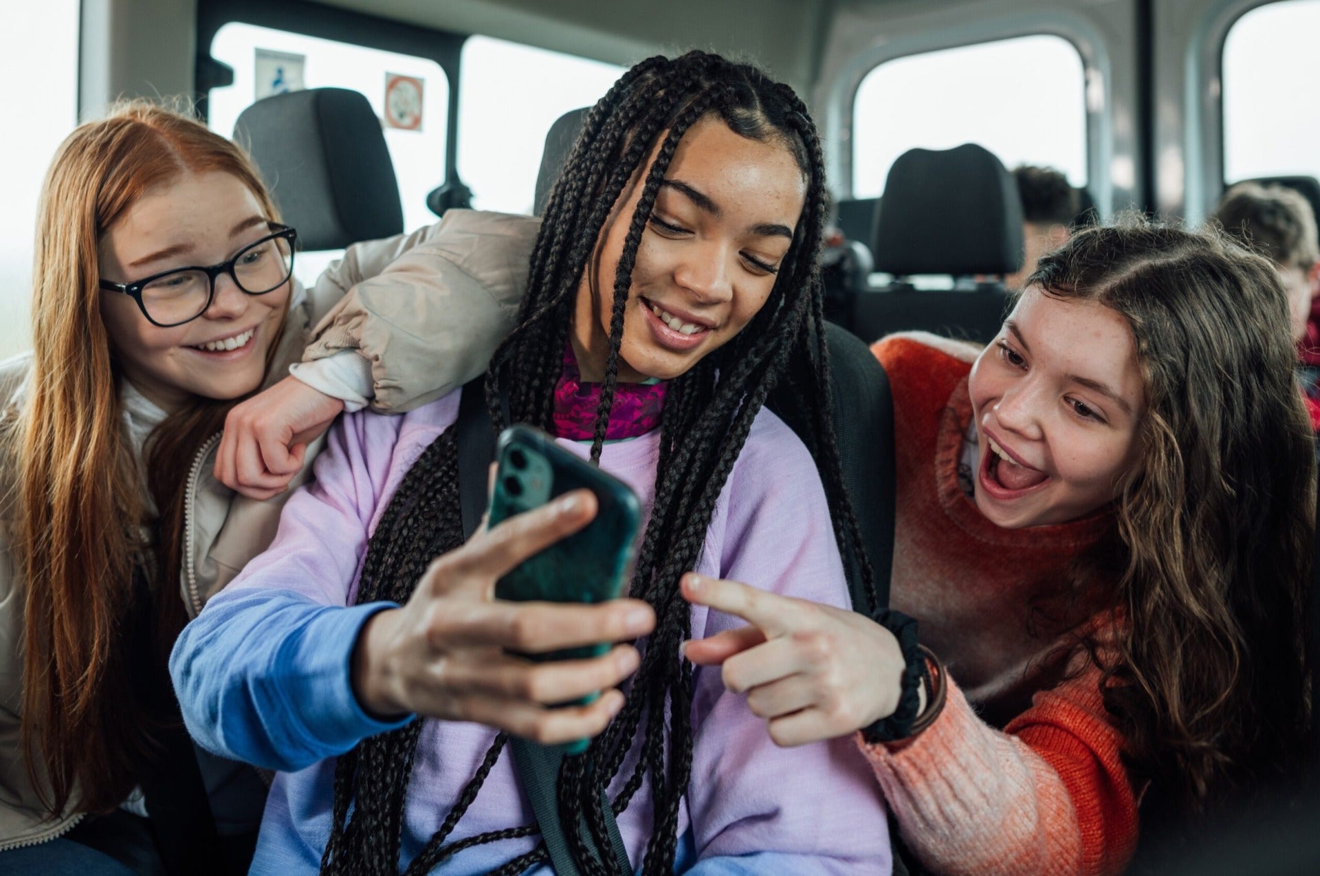 A photo of a group of three teenagers on the bus. They are looking a mobile phone and smiling and pointing at the screen.