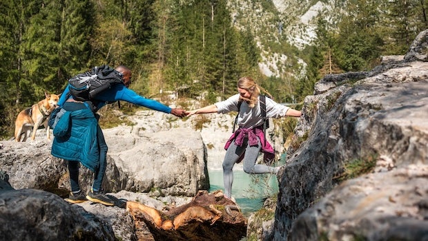 A person in hiking gear helps another person in hiking gear cross a river, representing trust-building in tech.